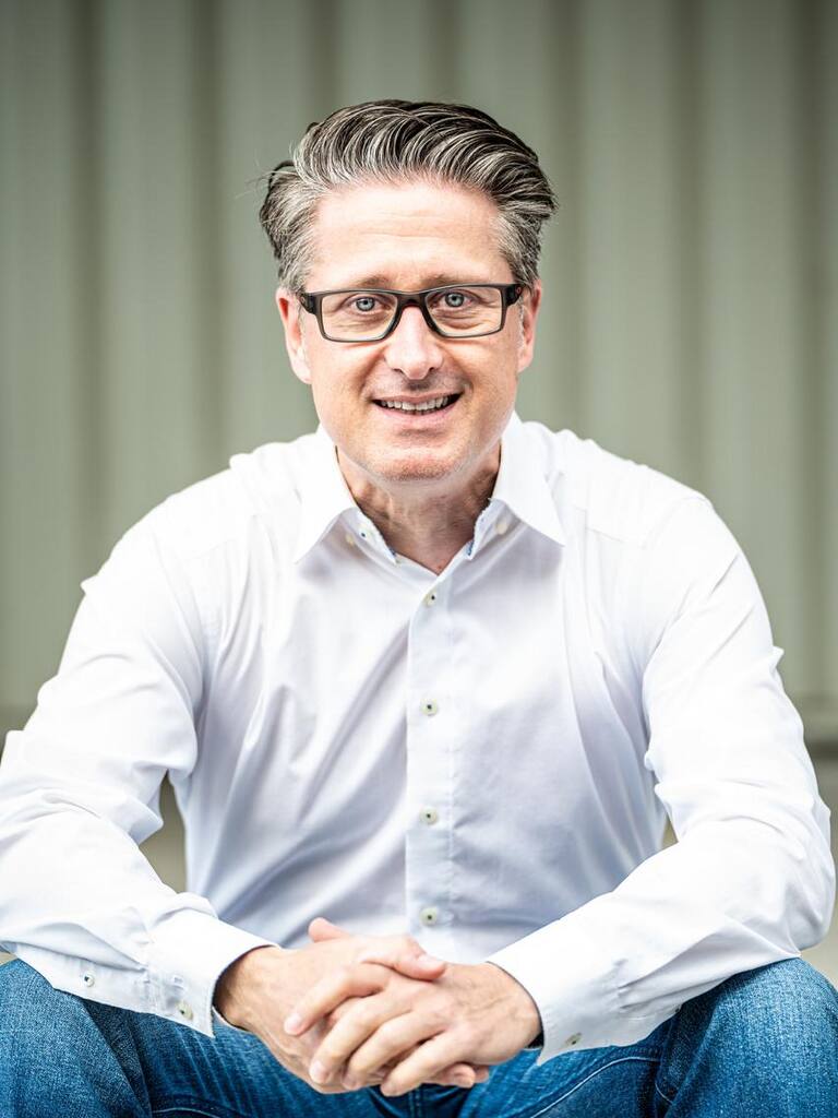 Michael Danner im weißen Hemd und Jeans, sitzend mit gefalteten Händen, vor einer grauen Wand